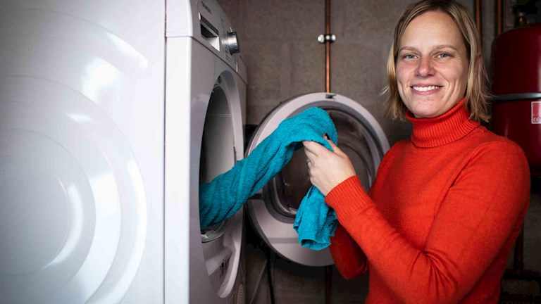 Aurélie se tient à côté de sa machine à laver et sort une serviette bleue
